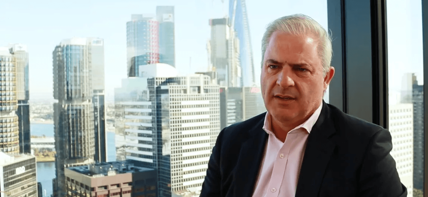 John Garret looking to the side, in a charcoal jacket and pink shirt, against a background of the Sydney skyline