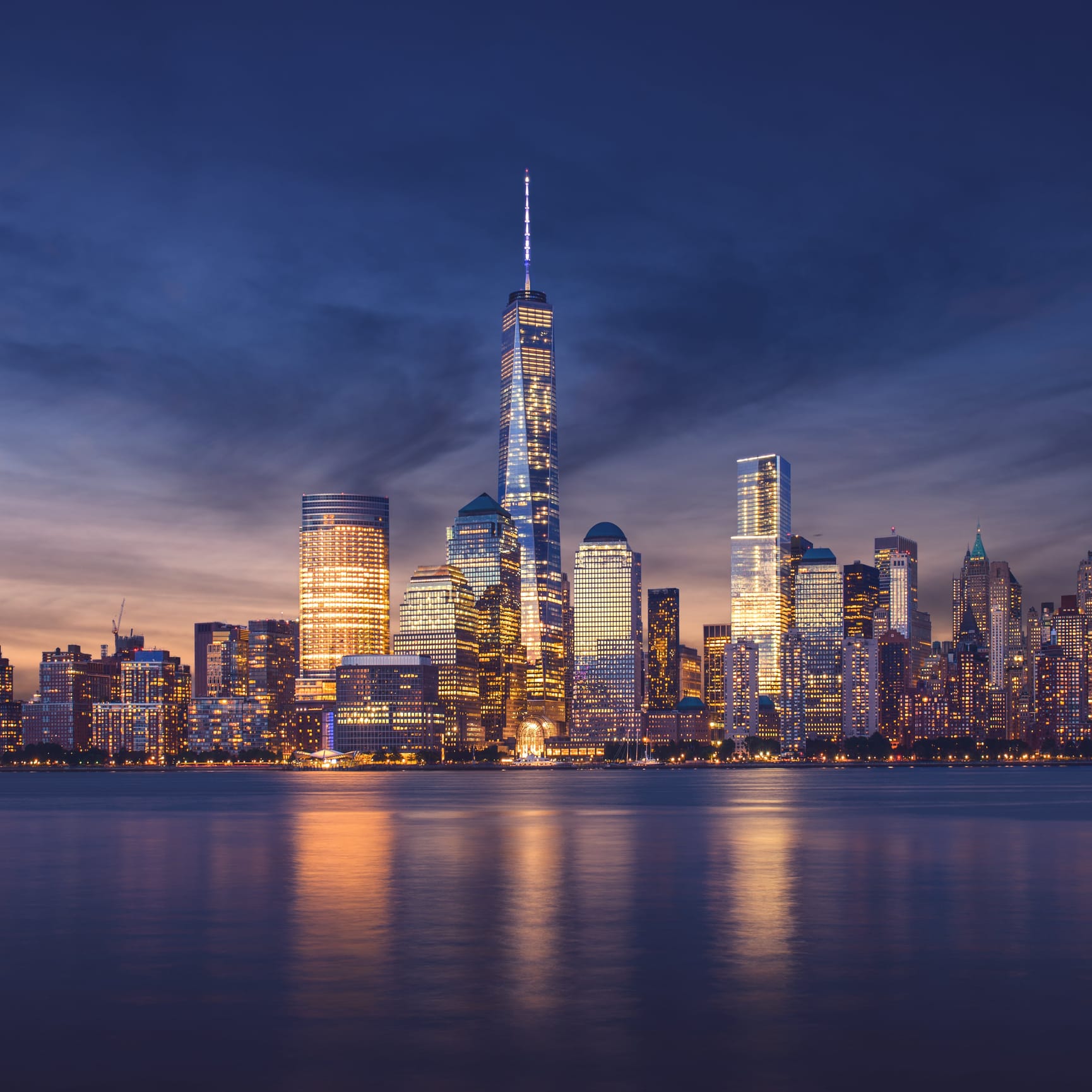 Evening photo of Manhattan with deep blue and purple tones with golden lights shimmering from office buildings.
